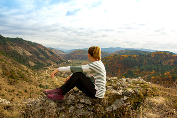 Naklejka na ściany i meble A woman sits on stones at the edge of a mountain peak and looks into the distance at the mountain landscape and the river flowing between the mountains.
