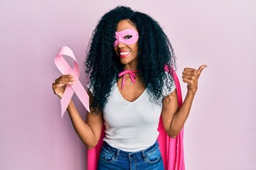 Middle age african american woman wearing super hero costume holding pink cancer ribbon pointing...