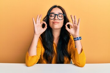Beautiful brunette young woman wearing glasses and casual clothes sitting on the table relax and smiling with eyes closed doing meditation gesture with fingers. yoga concept.