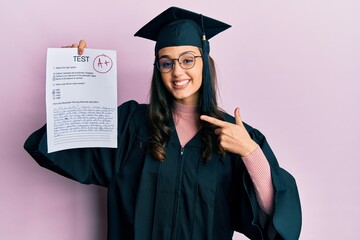 Young hispanic woman wearing graduation uniform holding passed exam smiling happy pointing with hand and finger