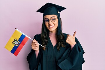 Young hispanic woman wearing graduation uniform holding ecuador flag smiling happy and positive, thumb up doing excellent and approval sign