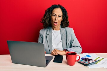 Beautiful middle age woman working at the office drinking a cup of coffee in shock face, looking...