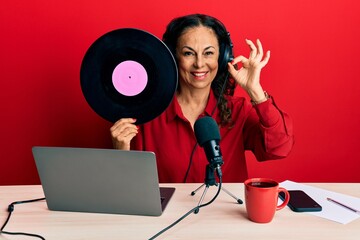 Beautiful middle age woman working at radio studio holding vinyl disc doing ok sign with fingers, smiling friendly gesturing excellent symbol
