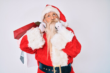 Old senior man with grey hair and long beard wearing santa claus costume holding shopping bags serious face thinking about question with hand on chin, thoughtful about confusing idea