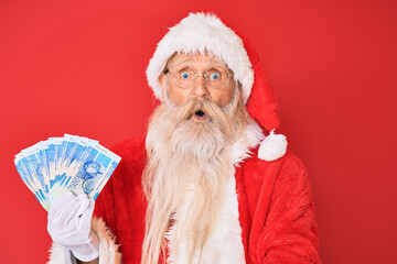 Old senior man with grey hair and long beard wearing santa claus costume holding african rands scared and amazed with open mouth for surprise, disbelief face