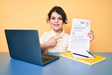 Cute hispanic child showing failed exam sitting on the desk smiling happy pointing with hand and finger