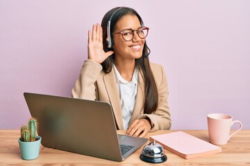 Beautiful hispanic woman working at the office wearing operator headset smiling with hand over ear listening and hearing to rumor or gossip. deafness concept.