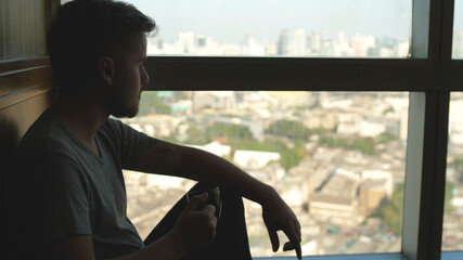 Portrait of young bearded man drinks cup of fresh hot coffee with steam and looks out window at dawn. Morning coffee concept