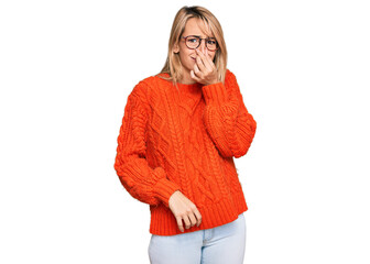 Beautiful blonde woman wearing casual clothes and glasses smelling something stinky and disgusting,...