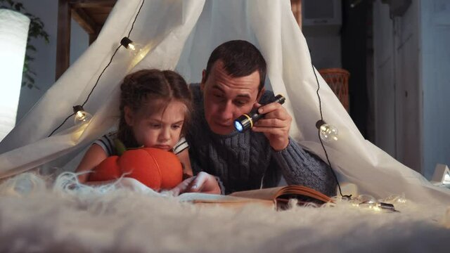 happy family at home dream. dad and daughter happy family at home reading lockdown a book in the evening in stay home homemade wigwam house. kid dream coronavirus concept. dad daughter read book