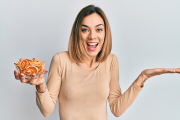 Young caucasian blonde woman holding bowl of dry orange celebrating achievement with happy smile and winner expression with raised hand