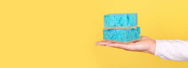 Hand with turquoise cleaning sponge on yellow background.