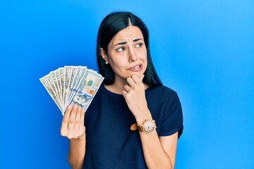 Beautiful young woman holding dollars thinking worried about a question, concerned and nervous with hand on chin