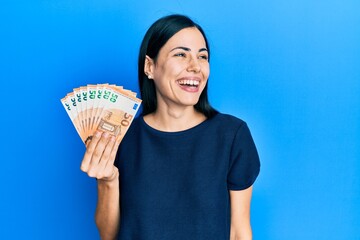 Beautiful young woman holding bunch of 50 euro banknotes looking away to side with smile on face, natural expression. laughing confident.
