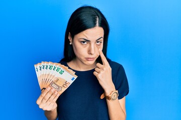 Beautiful young woman holding bunch of 50 euro banknotes pointing to the eye watching you gesture, suspicious expression