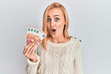 Beautiful caucasian blonde woman holding 10 euro banknotes scared and amazed with open mouth for...