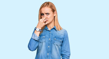 Beautiful caucasian woman wearing casual denim jacket smelling something stinky and disgusting, intolerable smell, holding breath with fingers on nose. bad smell