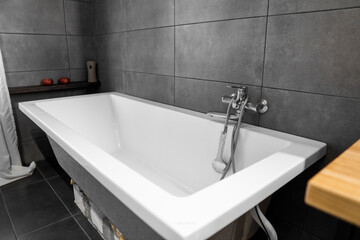 Bathroom interior with grey tile on a wall, stylish white bath with a silver tap. Minimalism in interior of bathroom.