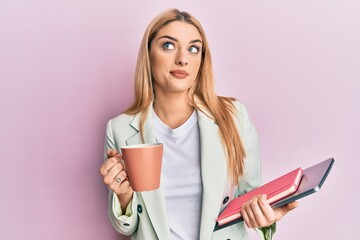 Young caucasian woman drinking a cup of coffee and holding laptop smiling looking to the side and staring away thinking.