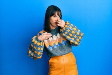 Young hispanic woman wearing casual winter sweater smelling something stinky and disgusting,...