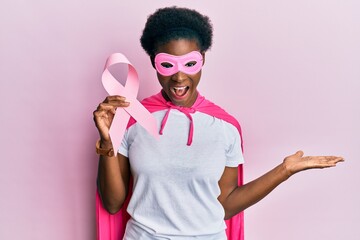 Young african american girl wearing superhero costume holding pink cancer ribbon celebrating...