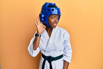 Young african american girl wearing taekwondo kimono and protection helmet smiling with hand over ear listening and hearing to rumor or gossip. deafness concept.