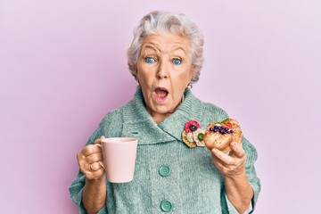 Senior grey-haired woman drinking a cup of coffee and eating bun afraid and shocked with surprise...