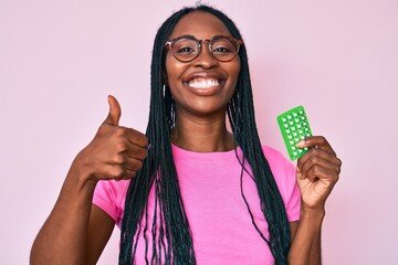 African american woman with braids holding birth control pills smiling happy and positive, thumb up doing excellent and approval sign