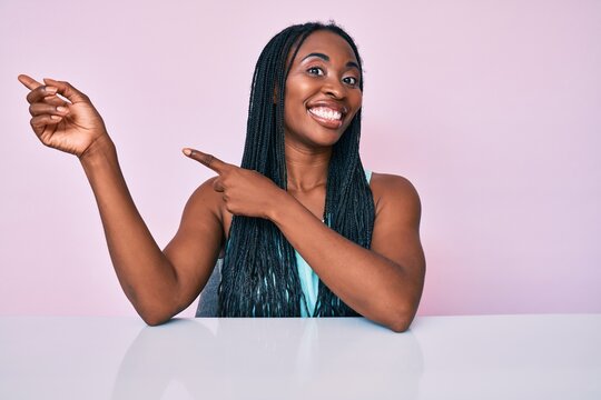 African american woman with braids wearing casual clothes sitting on the table smiling and looking at the camera pointing with two hands and fingers to the side.