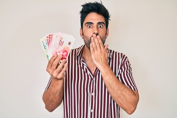 Handsome hispanic man with beard holding israel shekels covering mouth with hand, shocked and...