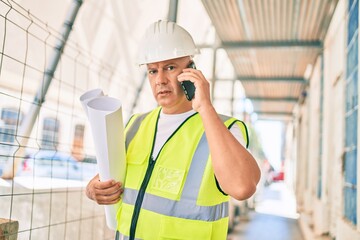 Middle age architect man talking on the smartphone and holding blueprints at the city.