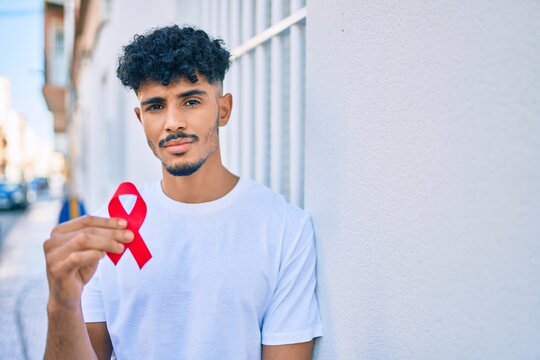 Young arab man with serious expression holding hiv awaraness red ribbon leaning on the wall.