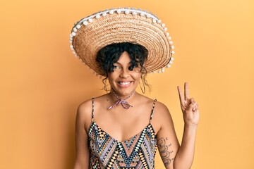 Young hispanic woman holding mexican hat smiling with happy face winking at the camera doing victory sign. number two.