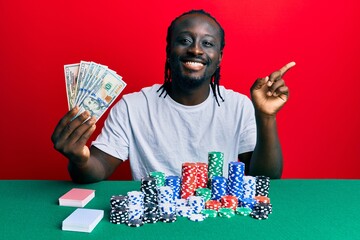 Handsome young black man playing poker holding 100 dollars banknotes smiling happy pointing with hand and finger to the side