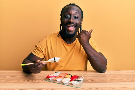 Handsome young black man eating sushi sitting on the table pointing thumb up to the side smiling happy with open mouth