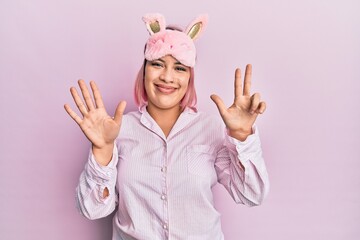 Hispanic woman with pink hair wearing sleep mask and pajama showing and pointing up with fingers number eight while smiling confident and happy.