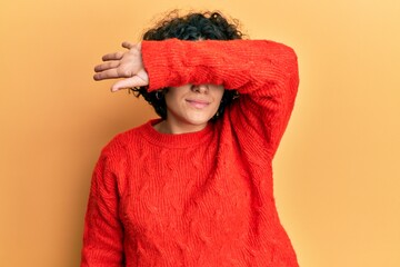 Young hispanic woman with curly hair wearing casual winter sweater covering eyes with arm, looking serious and sad. sightless, hiding and rejection concept