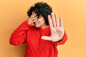 Fototapeta na wymiar Young hispanic woman with curly hair wearing casual winter sweater covering eyes with hands and doing stop gesture with sad and fear expression. embarrassed and negative concept.