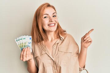 Young caucasian woman holding bunch of 5 euro banknotes smiling happy pointing with hand and finger to the side