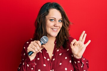 Young caucasian woman singing song using microphone doing ok sign with fingers, smiling friendly gesturing excellent symbol