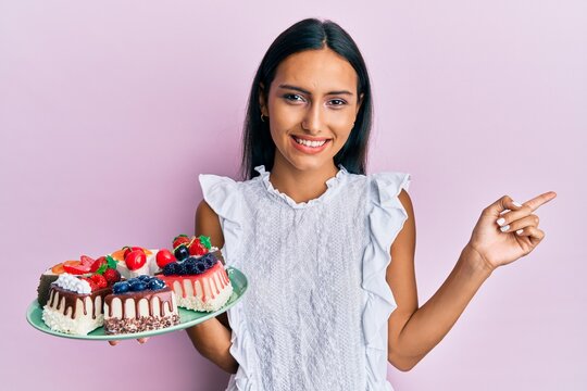 Young brunette woman holding cake slices smiling happy pointing with hand and finger to the side