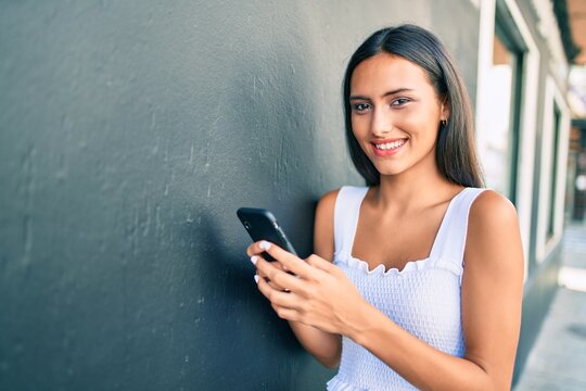 Young latin girl smiling happy using smartphone leaning on the wall.