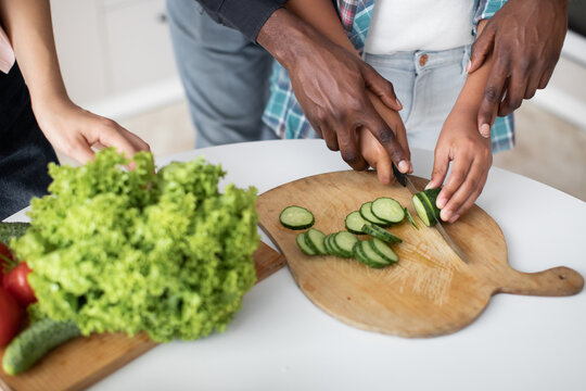 Close up cropped image of hands of black daddy teaching his teenage daughter holding knife and cut fresh cucumber. Healthy food with family lifestyle