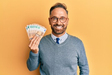 Handsome middle age man holding bunch of 50 euro banknotes looking positive and happy standing and smiling with a confident smile showing teeth - obrazy, fototapety, plakaty