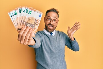 Handsome middle age man holding bunch of euros celebrating achievement with happy smile and winner expression with raised hand - obrazy, fototapety, plakaty