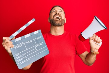 Handsome middle age man holding video film clapboard and megaphone angry and mad screaming...