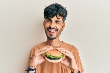 Young hispanic man eating a tasty classic burger winking looking at the camera with sexy...
