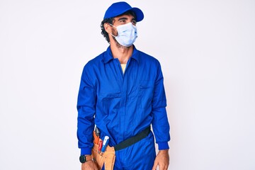 Handsome young man with curly hair and bear wearing handyman uniform and covid-19 safety mask looking to side, relax profile pose with natural face and confident smile.
