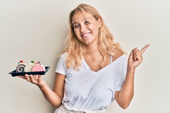 Young blonde girl holding cake sweets smiling happy pointing with hand and finger to the side