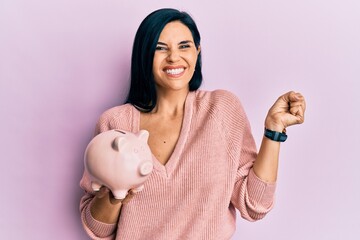 Young caucasian woman holding piggy bank screaming proud, celebrating victory and success very...
